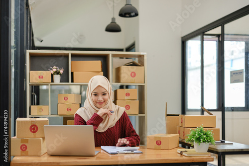 A portrait of a successful young Asian Muslim female online seller sits at her working desk with a laptop and cardboard boxes on a table.