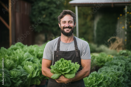 a smiling man with a beard and apron standing next to a bush of lettuce and smiling at the camera.