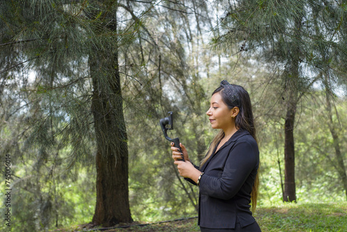 Woman vlogger in pine forest filming with cell phone on a stabilizer.