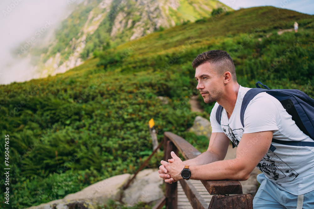 A man in light-colored clothes leans on a wooden fence. Against the background of mountains covered with vegetation. Hiking in the mountains in summer