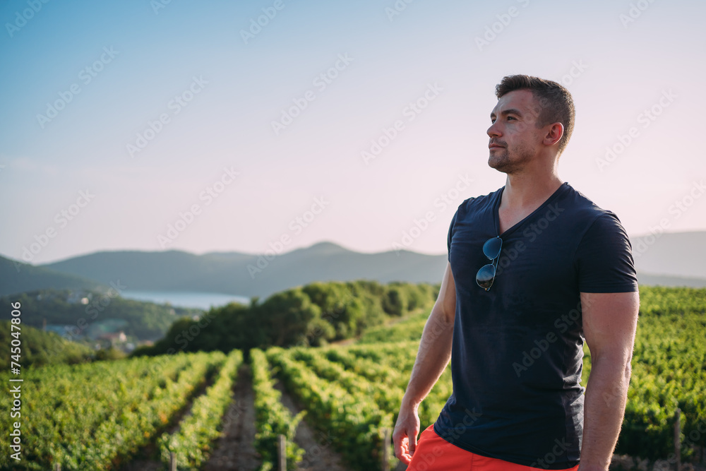 A man in a dark T-shirt stands on a vine alley in a vineyard. Mountains in the background, sunset. Vineyards at the winery