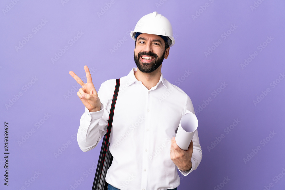 Young architect man with helmet and holding blueprints over isolated purple background smiling and showing victory sign