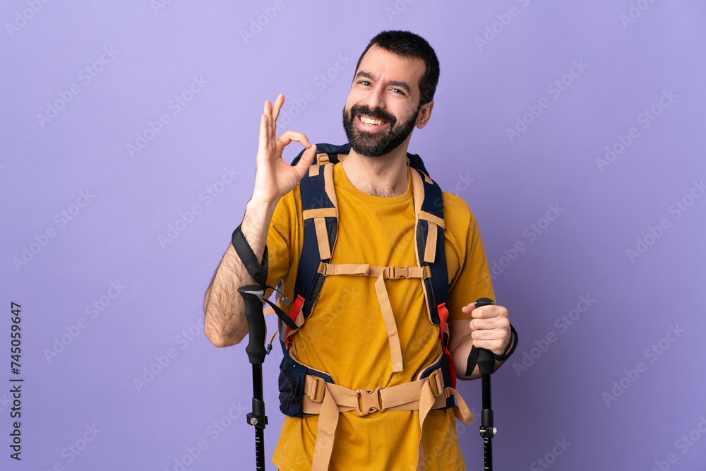 Caucasian handsome man with backpack and trekking poles over isolated background showing ok sign with fingers Caucasian handsome man with backpack and trekking poles over isolated background showing ok sign with fingers