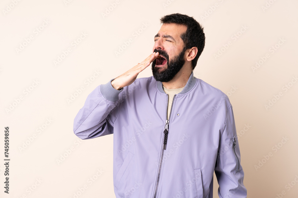 Caucasian man with beard wearing a jacket over isolated background yawning and covering wide open mouth with hand Caucasian man with beard wearing a jacket over isolated background yawning and covering wide open mouth with hand