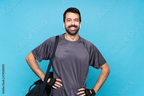 Young sport man with beard over isolated blue background posing with arms at hip and smiling