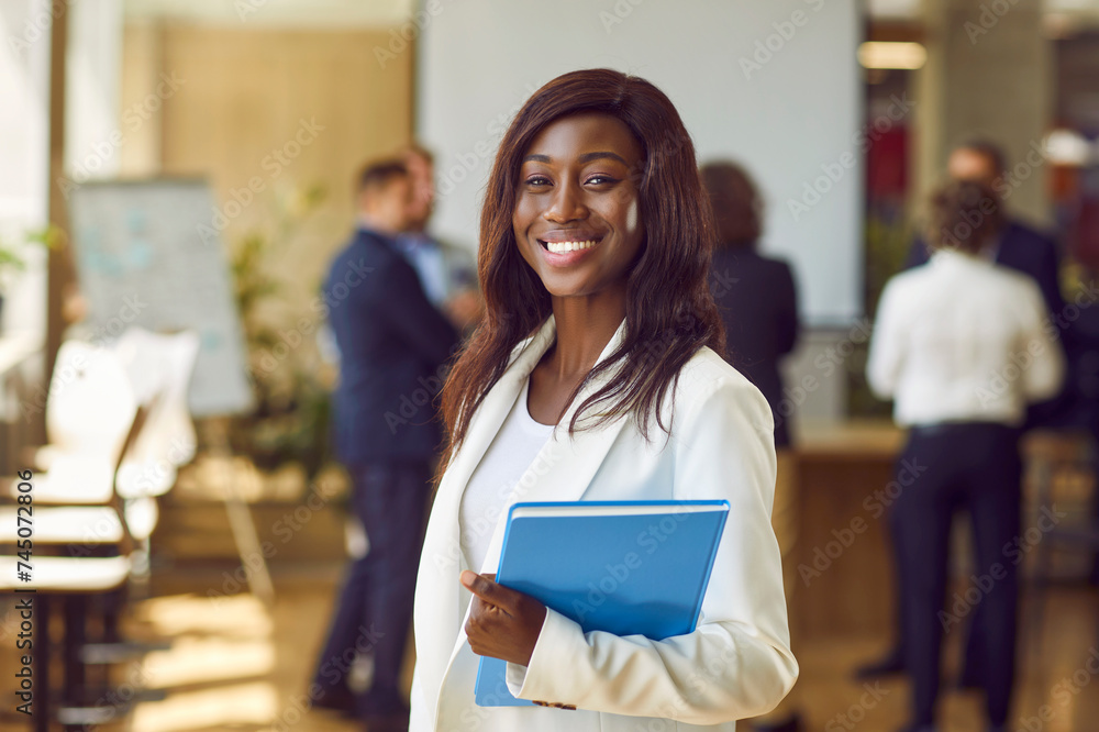 Fototapeta premium Close up portrait of a happy smiling confident african american business woman leader proudly looking at the camera with a team of company employees talking in the background in office.