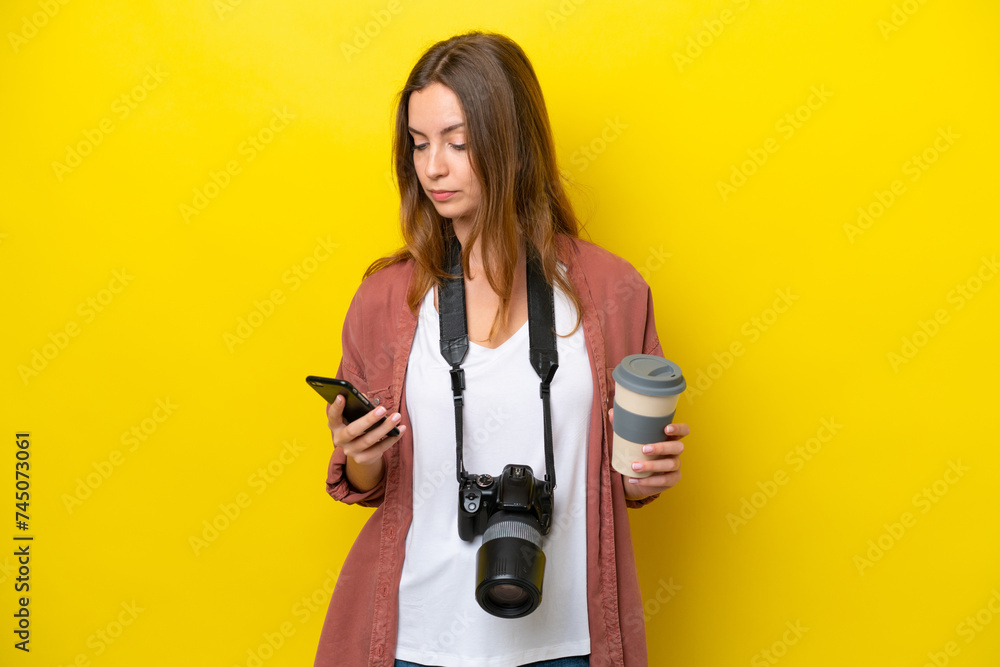 Young photographer caucasian woman isolated on yellow background holding coffee to take away and a mobile Young photographer caucasian woman isolated on yellow background holding coffee to take away and a mobile