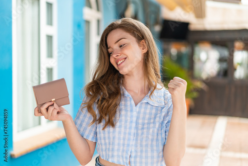 Young redhead woman holding a wallet at outdoors celebrating a victory