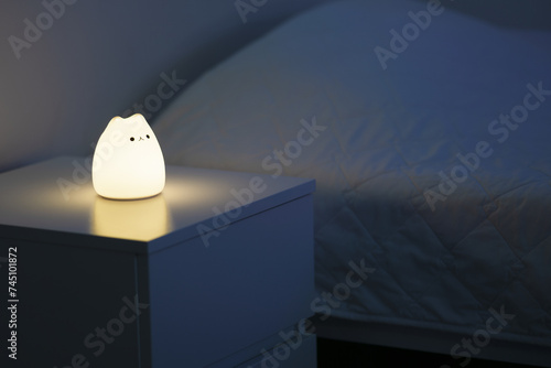 Cute cat shaped night lamp standing on a bedside table next to bed. Bedside lamp. Night lamp standing next to bed. Bedroom lamp on a night table next to a sleeping bed in a dark room. 