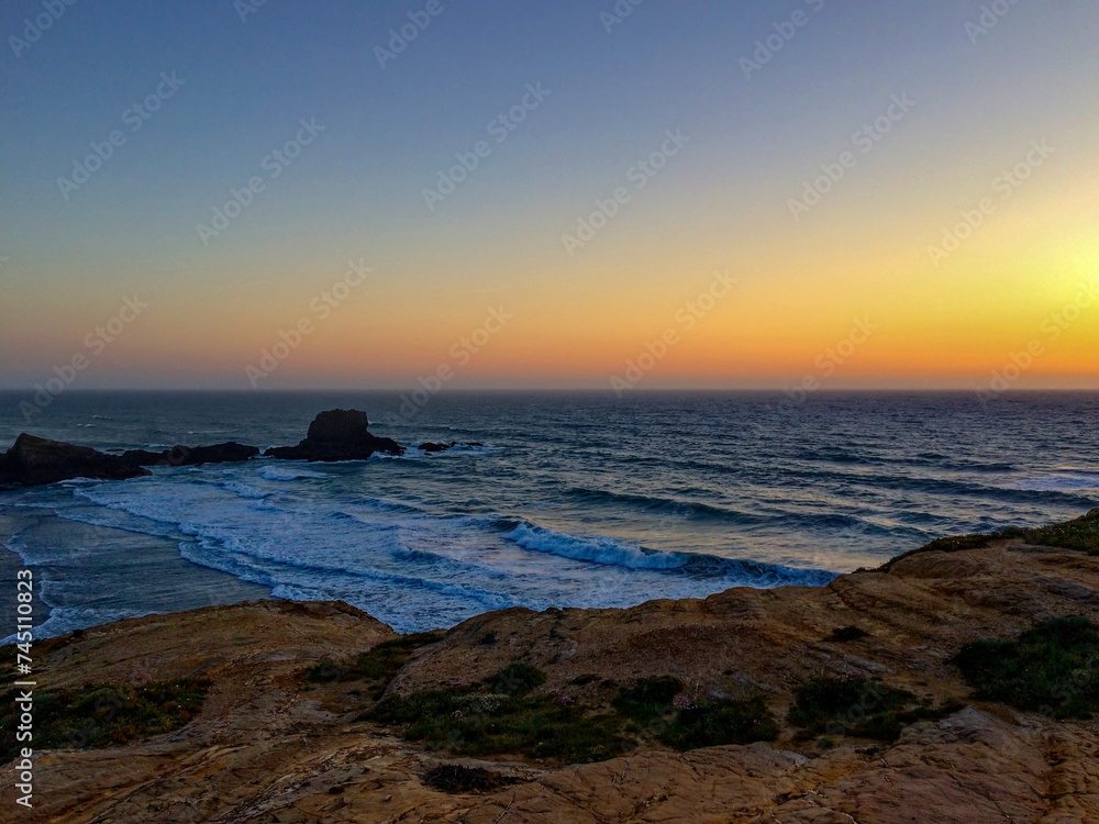 The image captures a beautiful sunset over a rugged coastline with waves crashing against the rocks.