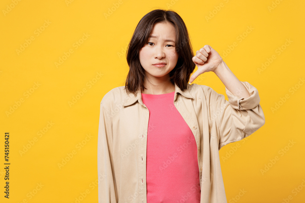 Young dissatisfied sad woman of Asian ethnicity wear pink t-shirt beige shirt pastel casual clothes showing thumb down dislike gesture isolated on plain yellow background studio. Lifestyle portrait. Young dissatisfied sad woman of Asian ethnicity wear pink t-shirt beige shirt pastel casual clothes showing thumb down dislike gesture isolated on plain yellow background studio. Lifestyle portrait.