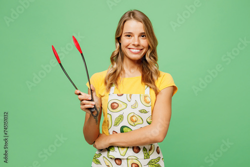 Young housewife housekeeper chef cook baker woman wear apron yellow t-shirt hold kitchen salad serving plastic pair of tongs for grill isolated on plain pastel green background. Cooking food concept. photo