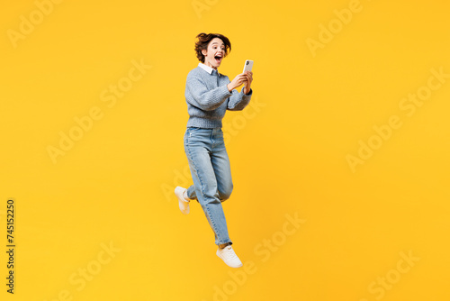 Full body excited young woman she wears grey knitted sweater shirt casual clothes jump high hold in hand use mobile cell phone isolated on plain yellow background studio portrait. Lifestyle concept.