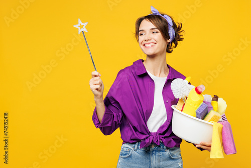 Young smiling woman wear purple shirt hold basin with detergent bottles do housework tidy up hold magic wand fairy stick look aside on area isolated on plain yellow background. Housekeeping concept.