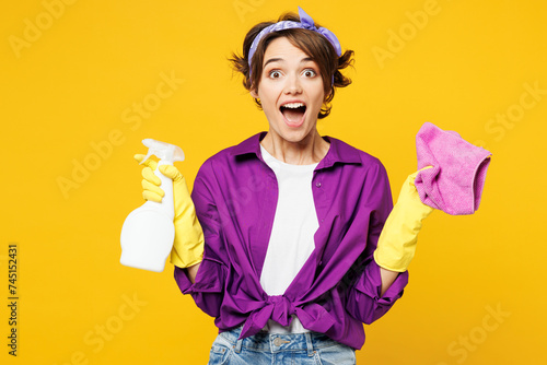 Young surprised shocked woman wears purple shirt rubber gloves do housework tidy up hold in hand rag spray bottle look camera isolated on plain yellow background studio portrait. Housekeeping concept. photo