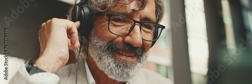 Panorama, Close up portrait of mature businessman with beard in eyeglasses and headphones, sits in an outdoor cafe and works using tablet