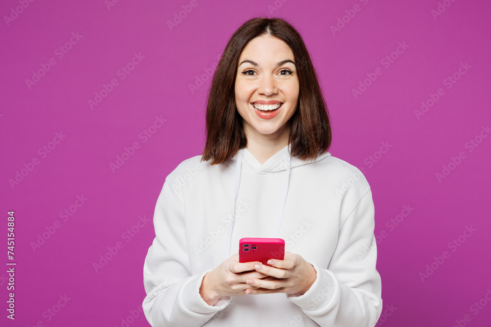 Young smiling happy Caucasian woman she wear light hoody casual clothes hold in hand use mobile cell phone chatting online isolated on plain purple color background studio portrait. Lifestyle concept. Young smiling happy Caucasian woman she wear light hoody casual clothes hold in hand use mobile cell phone chatting online isolated on plain purple color background studio portrait. Lifestyle concept.