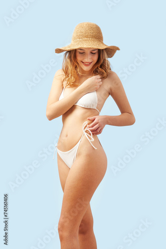 Smiling woman wearing a sun hat, fastening her white bikini, is isolated a light blue background. Banner or poster image for the concept of a seaside holiday or shopping for a summer beach holiday
