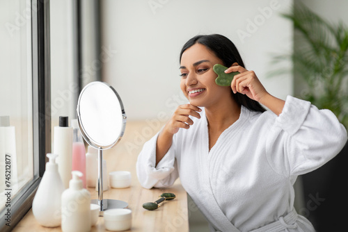 Smiling indian woman using gua sha, looking at mirror photo