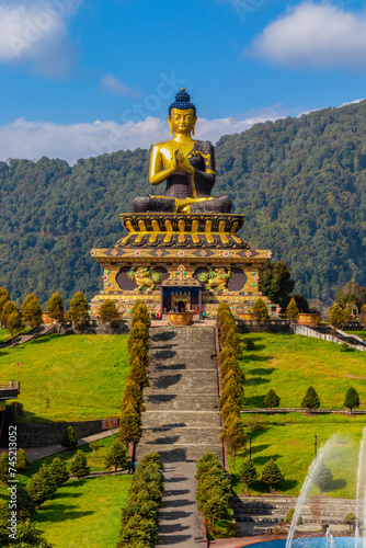 A series of steps leads towards a statue of Buddha located on the top of the mountain. The golden statue looks mesmerizing with a backdrop of a clear blue sky with a Cloudscape. Ravangla, Sikkim, Ind