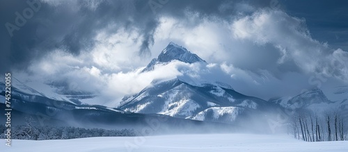A grand mountain rises above a snow-covered landscape, with its peak and slopes blanketed in pristine white snow. The scene is enhanced by the dramatic backdrop of thick, heavy clouds hovering above.
