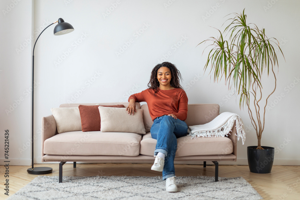 Cheerful young black woman sitting comfortably on sofa with cushions at home Cheerful young black woman sitting comfortably on sofa with cushions at home