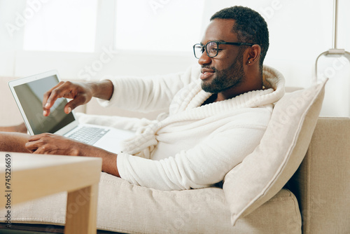 African American freelancer working on a laptop in a modern home The young man dressed in a bathrobe sits on a comfortable sofa in the living room, typing with a smile on his face It's a sunny