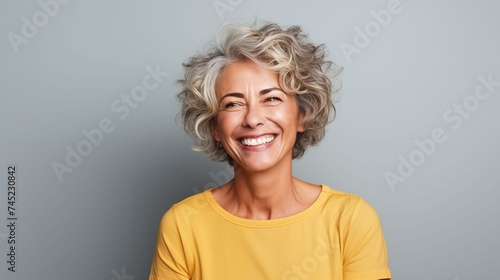 Close up of  mature woman with white skin, grey short hair, wavy hair and a clear yellow t shirt, isolated in a light grey studio. Portrait person.