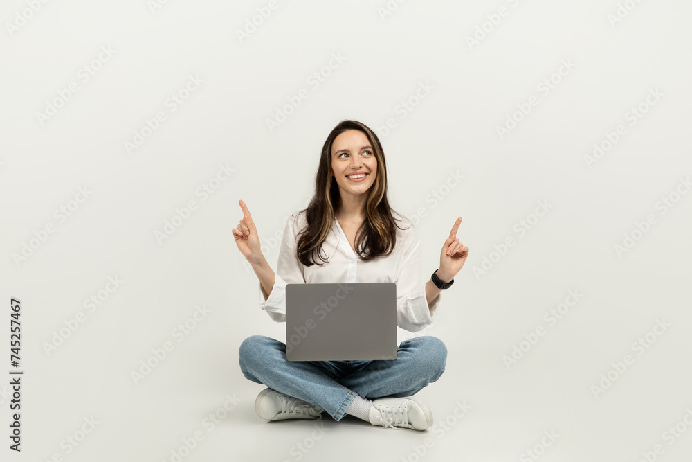Happy woman sitting cross-legged on the floor with a laptop, pointing upwards Happy woman sitting cross-legged on the floor with a laptop, pointing upwards