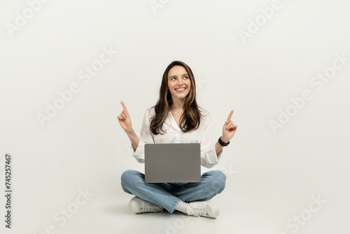 Happy woman sitting cross-legged on the floor with a laptop, pointing upwards © Prostock-studio