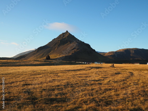 Beautiful view of Mount Stapafell and Bárður Snæfellsás the mythical protector, grassland in golden sunlight in the foreground, with a couple of travelers far away, in Snaefellsnes Peninsula of Icelan
