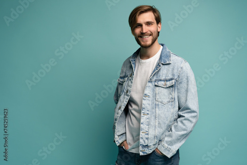 Portrait of stylish brutal young bearded European man wearing jeans jacket, posing at blue blank wall with copy space for your text or promotional content