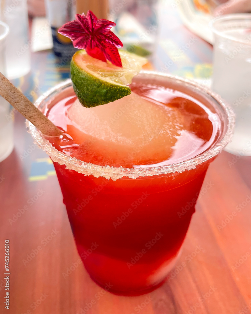 Frozen strawberry daiquiri sitting on table, with its rim salted and a vibrant lime wedge adds a zesty pop of color. Completing the picturesque scene is a tropical flower resting on top the drink.