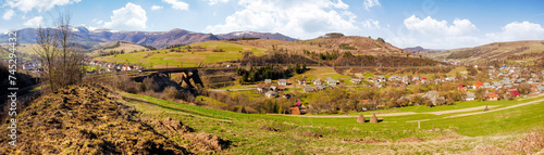 panorama of mountainous rural landscape with village in the valley. carpathian countryside scenery with arable on hills in front of borzhava ridge with snow capped top on a sunny morning in spring