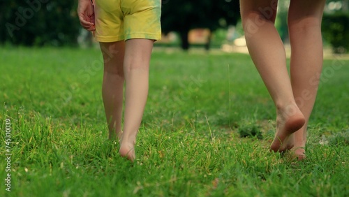 Barefoot mom, boy, girl go on green grass in park. Bare feet mom, child, close-up go on green grass. Concept healthy lifestyle. Family walk barefoot on green lawn on sunny day. Kid, mother fun outdoor