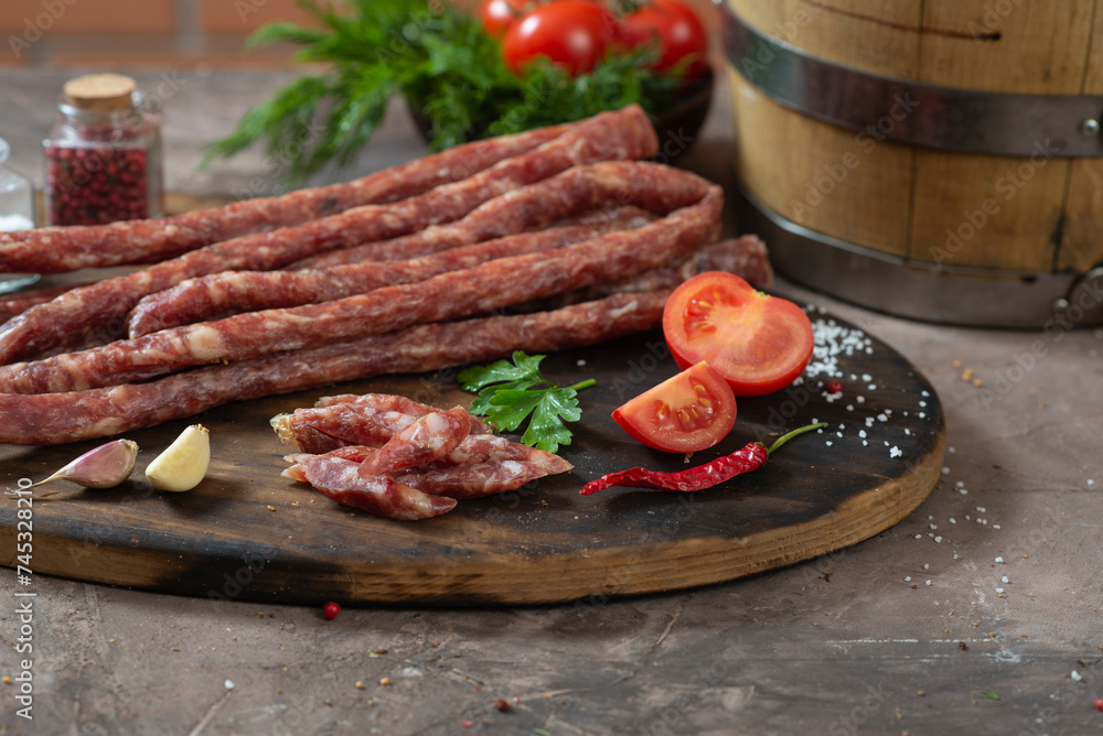 Rustic dry thin sausages among vegetables and herbs on a dark table Rustic dry thin sausages among vegetables and herbs on a dark table