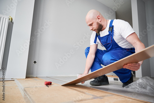 Male construction worker laying laminate wooden plank on floor underlayment in apartment under renovation. Man in workwear installing laminate timber flooring in living room. Home renovation concept.