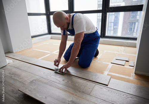 Man in work overalls using metal construction ruler and pen while drawing line on laminate flooring board. Male floor installer preparing laminate plank for installation in apartment under renovation.