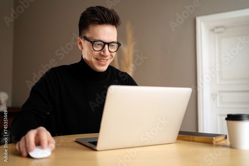 A young man with glasses smiles while working on a laptop, a coffee cup and notebook beside him.