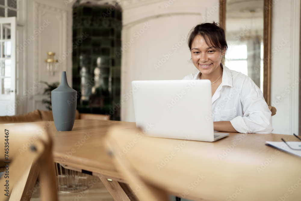 A smiling woman in a white blouse works on her laptop at a wooden table in a light-filled room with elegant interior. A smiling woman in a white blouse works on her laptop at a wooden table in a light-filled room with elegant interior.