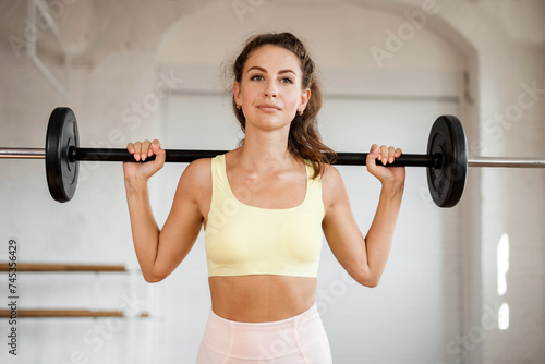 Confident young woman lifting a barbell in a bright gym, showcasing strength and focus.