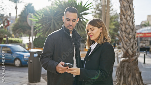 A man and woman engaging with a smartphone while standing together outdoors on a sunny city street.
