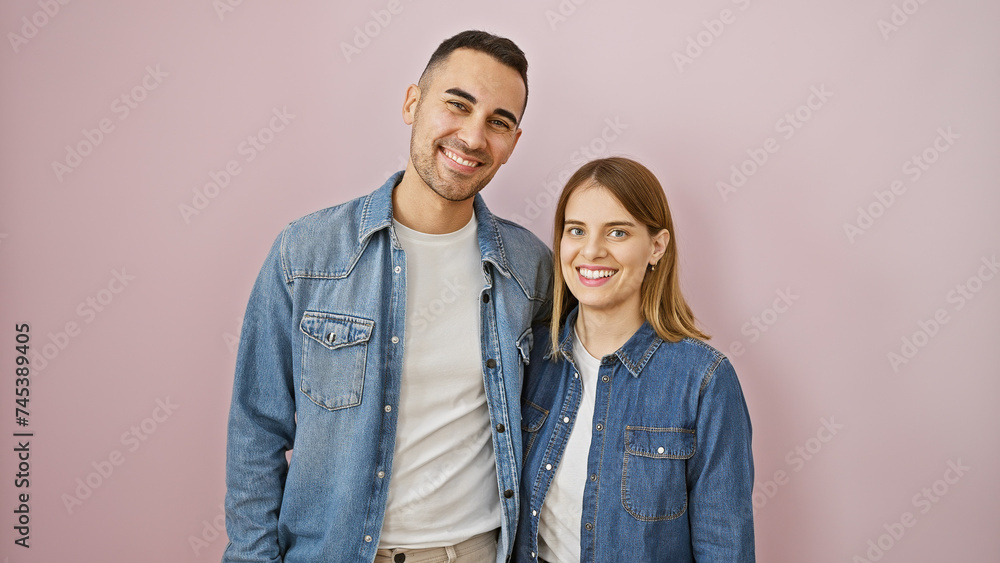 A smiling man and woman in denim stand together against a plain pink wall, depicting a happy couple. A smiling man and woman in denim stand together against a plain pink wall, depicting a happy couple.