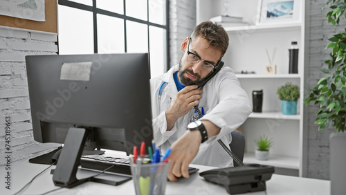 Young, handsome hispanic male doctor concentrated on diagnosis, talking on telephone, working online on computer at his indoor clinic workplace