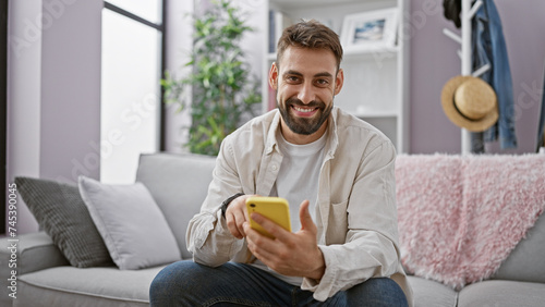 Handsome young hispanic man happily enjoying texting and surfing the internet on his smartphone, sitting cheerfully on the indoor sofa at his cosy home living room.