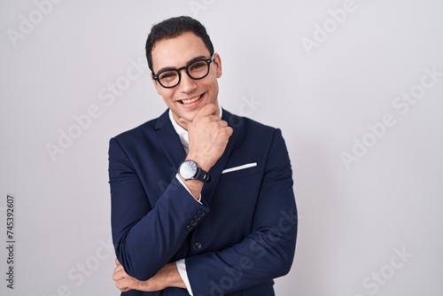 Young hispanic man wearing suit and tie looking confident at the camera smiling with crossed arms and hand raised on chin. thinking positive.