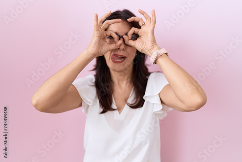 Middle age hispanic woman wearing casual white t shirt and glasses doing ok gesture like binoculars sticking tongue out, eyes looking through fingers. crazy expression. © Krakenimages.com