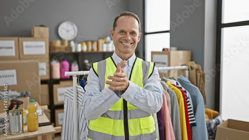 Confident smiling middle age man joyfully clapping hands, volunteering at charity center. handsome adult dons reflective vest, standing amidst donations, oozing unity, altruism in community. photo