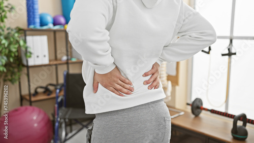 A young adult man in a physiotherapy clinic grimaces with back pain, indicating a need for rehab treatment.