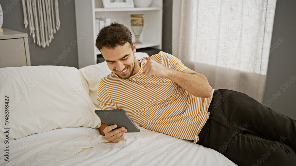 A cheerful young man with a beard enjoys using a tablet while relaxing in a modern bedroom.